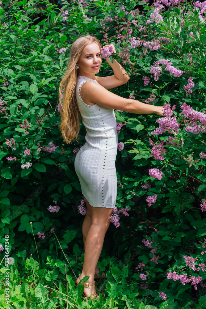 Portrait of a charming blond woman wearing beautiful white dress standing next to lilac bush. Portrait of a charming blond woman wearing beautiful white dress standing next to lilac bush.