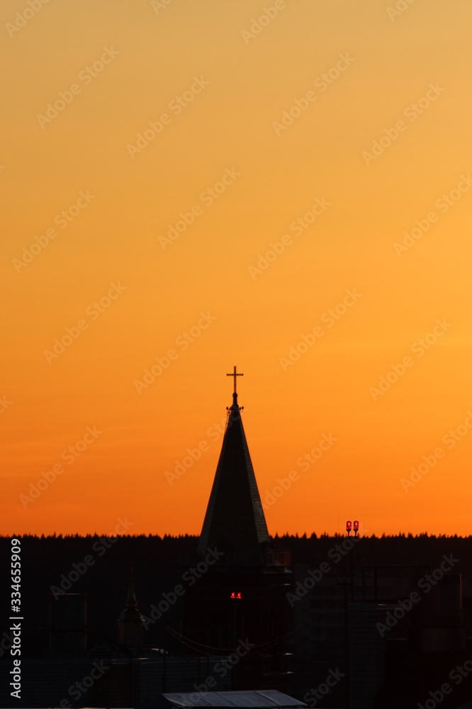 Obraz premium Sun below the horizon and the roof of the church with a cross on the background fiery dramatic orange sky at sunset or dawn backlit by the sun. Place for text and design.