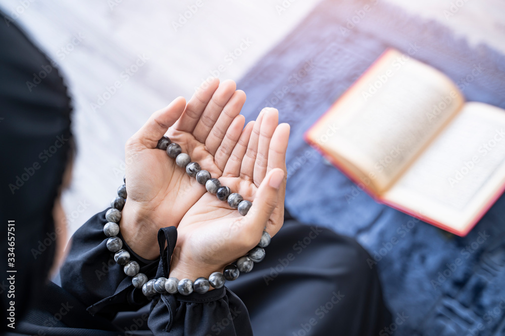 Close up beautiful asian muslim woman putting hands up to chest praying to allah with misbaha praying bead in hands sitting on knees on praying mat with quran resting on stand, bright light sunshine