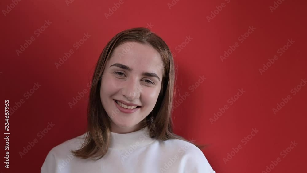 The girl smiles and laughs. Beautiful young woman in a white T-shirt smiles, shows good emotions. On a red background. Close-up face.