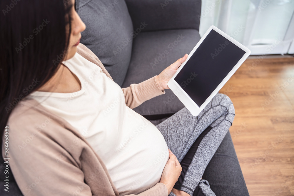 beautiful asian pregnant woman smiling using smart tablet device watching video placing hands on baby lump, sitting on sofa relaxing resting in living room with brick texture wall and white curtains beautiful asian pregnant woman smiling using smart tablet device watching video placing hands on baby lump, sitting on sofa relaxing resting in living room with brick texture wall and white curtains