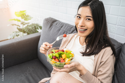 Beautiful asian pregnant woman holding a bowl of salad of vegetables and fruits  using a fork eating tomato. Eating healthy having lunch sitting on the grey sofa  relaxing and resting living room