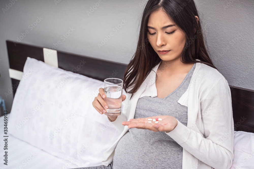 beautiful asian pregnant woman taking a medicine pill of vitamin and a glass of water sitting on white bed, relaxing and resting in a bed room as motherhood, wearing stretcher pants and white cardigan beautiful asian pregnant woman taking a medicine pill of vitamin and a glass of water sitting on white bed, relaxing and resting in a bed room as motherhood, wearing stretcher pants and white cardigan
