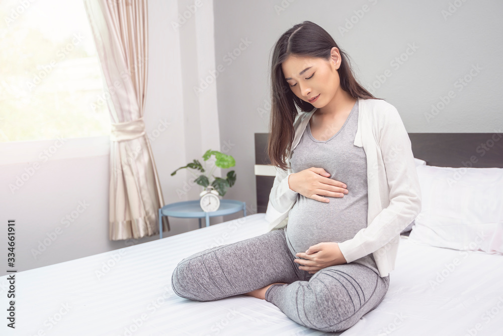 beautiful asian pregnant woman smiling and hands rubbing firmly on her baby stomach, joyfully and happiness, sitting on the white bed relaxing and resting in the bedroom with light shining from window beautiful asian pregnant woman smiling and hands rubbing firmly on her baby stomach, joyfully and happiness, sitting on the white bed relaxing and resting in the bedroom with light shining from window