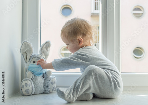 Easter 2021 concept with child in home quarantine playing at the window with his sick plush bunny wearing a medical mask against viruses during coronavirus COVID-2019 and flu outbreak.