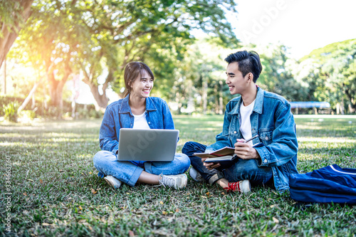 asian student couple studying outside on the grass, sitting down on the school campus using laptop computer, books, pencil and note book, studying hard under the trees nature in the evening sunlight