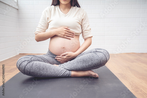 Body shot of pregnant woman caressing her baby belly, sitting on black yoga mat training yoga meditation, healthy and fit motherhood feeling happy and strong, pregnancy expecting future new born