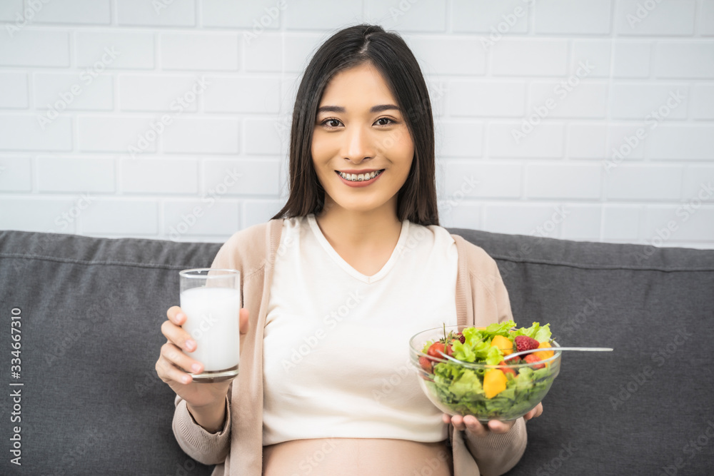 Beautiful asian pregnant woman holding a bowl of salad of vegetables and fruits, holding drinking glass of milk. Eating healthy having lunch sitting on the grey sofa, relaxing and resting living room Beautiful asian pregnant woman holding a bowl of salad of vegetables and fruits, holding drinking glass of milk. Eating healthy having lunch sitting on the grey sofa, relaxing and resting living room