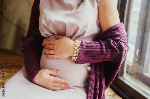 stylish pregnant in a delicate pink powder dress and a knitted purple maroon jumper at a vintage wooden window is stroking her big tummy and enjoying motherhood.