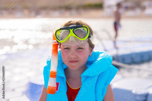 There is a portrait of a beautiful little girl in a life jacket. A happy child in a blue inflatable vest smiles cheerfully. Safe bathing for the child. Children's portrait in a mask for swimming