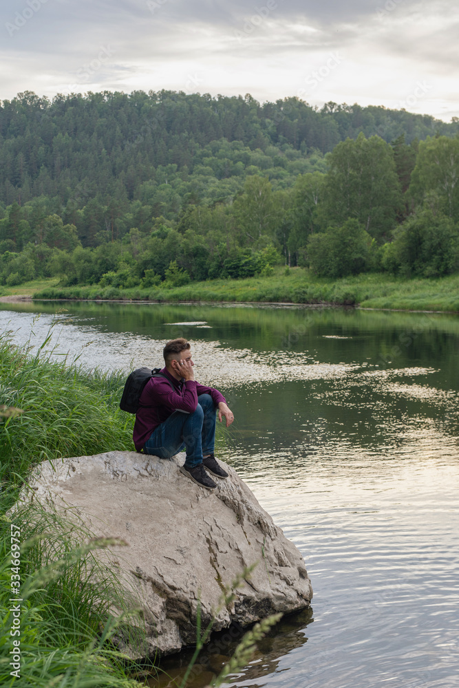 Side view portrait of young man tourist sitting on big stone on narrow forest river bank looking thoughtful and tired. Tourism, hiking, active leisure
