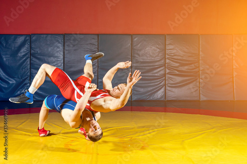 Two greco-roman wrestlers in red and blue uniform wrestling on a yellow wrestling carpet in the gym