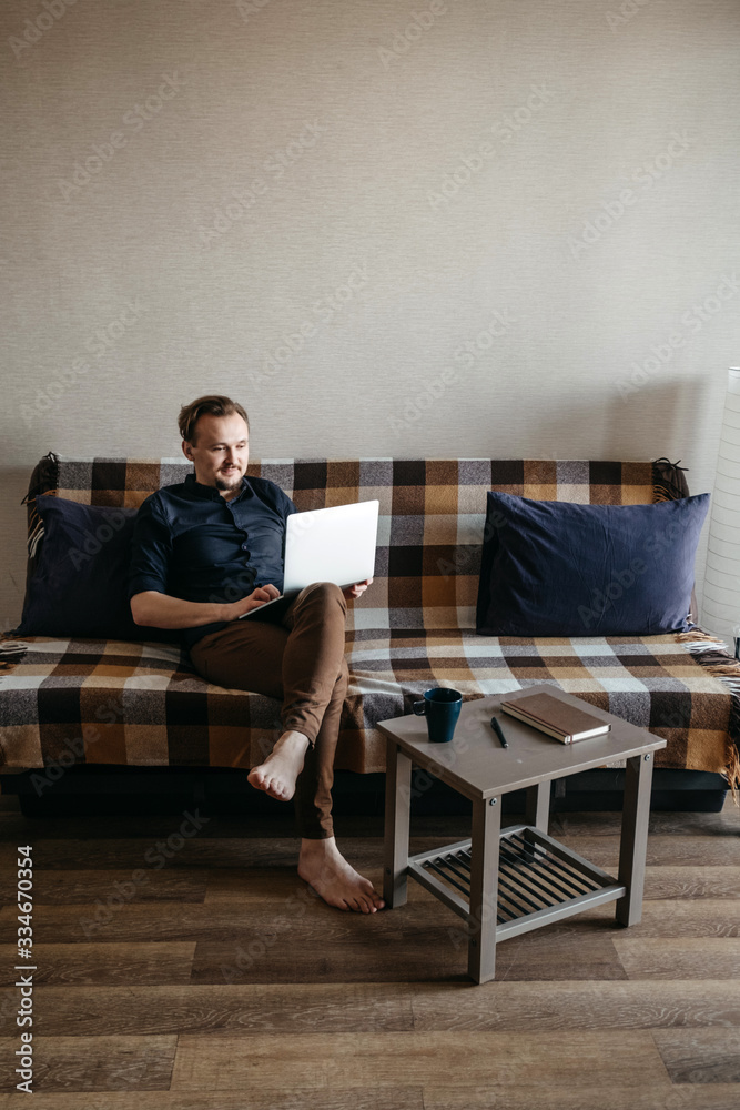 A young European guy in a dark blue shirt and brown trousers with a dark beard looks at a laptop. Sits on the couch, laptop on his knees. Home insulation. Home Office. A young European guy in a dark blue shirt and brown trousers with a dark beard looks at a laptop. Sits on the couch, laptop on his knees. Home insulation. Home Office.