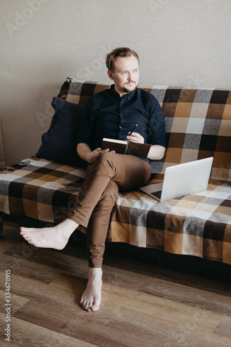 A young European guy in a dark blue shirt and brown trousers with a dark beard makes notes in a brown notebook. Sitting on the couch. On the sofa is a silver laptop. Home insulation. Home Office.