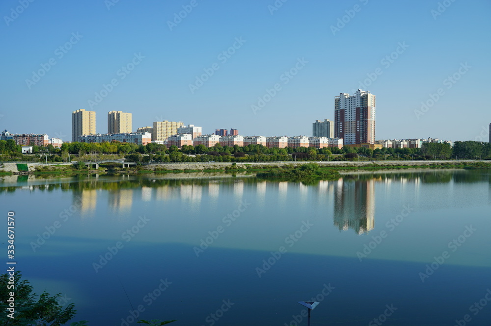 Cityscape by the river under blue sky, buildings reflecting on peaceful water