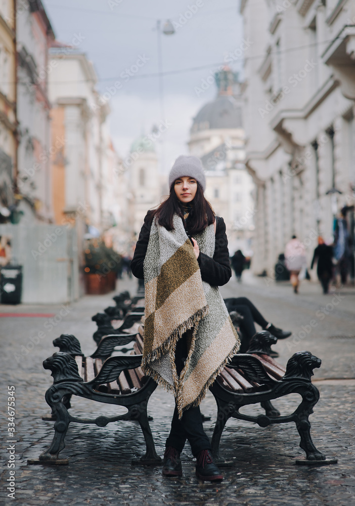A young caucasian girl in coat, angora hat, wide scarf is standing alone leaning on a cast-iron bench awaiting a date. Lviv, Ukraine.