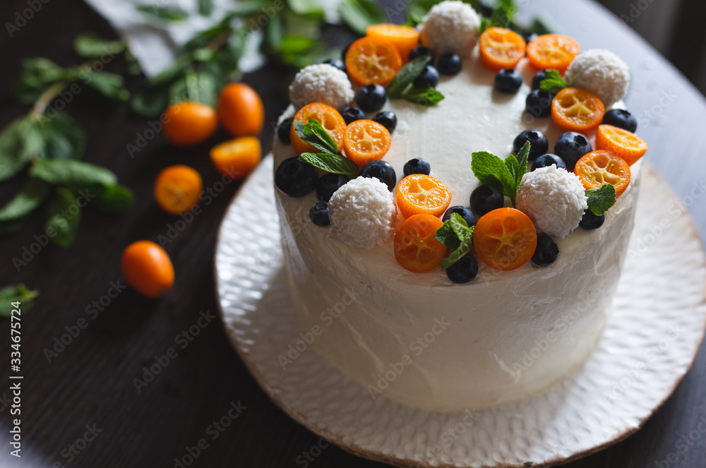 A celebratory fruit cake with whipped curd cream icing decorated with cut kumquats, blueberries, coconut sweets and mint leaves on a dark background. Selective focus