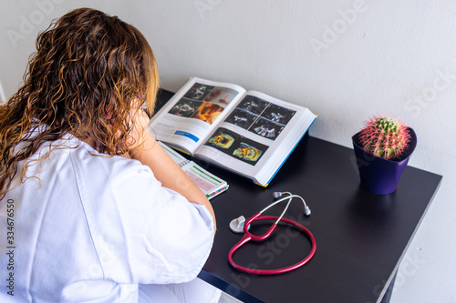 Young woman studying medicine sitting at a desk. There is also a cardiology textbook where you can see electrocardiograms.  On the desk there is a stethoscope and a cactus.  photo