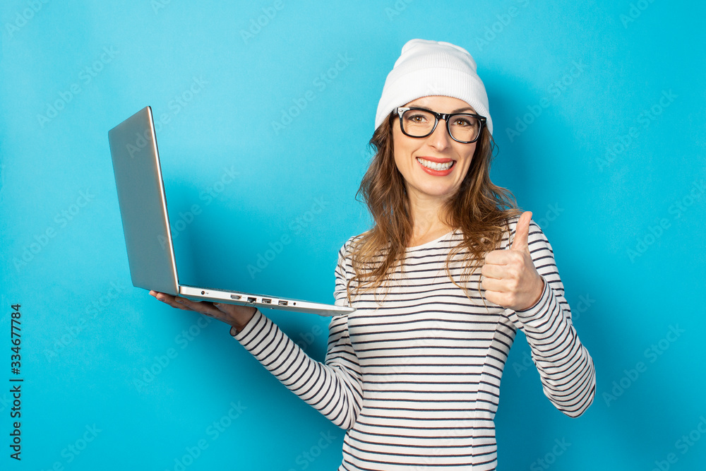 smiling young girl holds laptop, shows class hand gesture. Girl in glasses and a white hat on a blue background. Concept of work, freelance smiling young girl holds laptop, shows class hand gesture. Girl in glasses and a white hat on a blue background. Concept of work, freelance