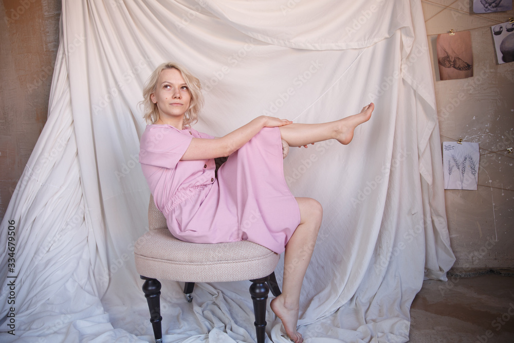 Fototapeta premium Full length photo of gorgeous woman 20s in pink dress sitting in soft armchair with legs crossed and seductively holding finger at lips isolated over blue background