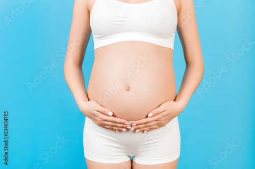 Cropped image of pregnant woman in white underwear touching her belly at blue background. Future mother is waiting her baby. Copy space © sosiukin