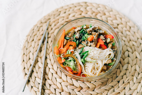 Japanese ramen soup in a glass plate with vegan ingredients  fresh vegetables in small plates  rice noodles  mushrooms and miso paste broth  on a natural wicker napkin on a white table