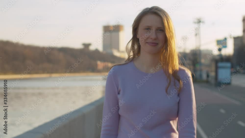 Portrait of a young smiling attractive woman walking on the embankment near the river against the background of the city. Blond woman with long hair outdoors