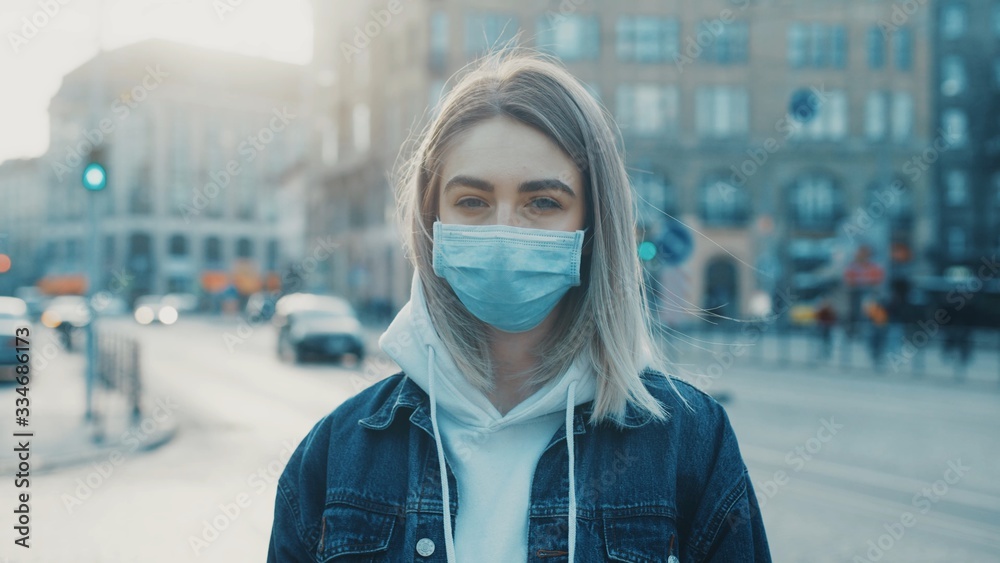 Young woman in medical mask standing in city street looking to camera road with cars on background Concept of health and safety life COVID-19 coronavirus virus protection pandemic in world Young woman in medical mask standing in city street looking to camera road with cars on background Concept of health and safety life COVID-19 coronavirus virus protection pandemic in world