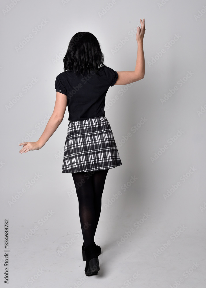 Portrait of a goth girl with dark hair wearing blue and plaid skirt with boots. Full length standing pose with back to the camera on a studio background. Portrait of a goth girl with dark hair wearing blue and plaid skirt with boots. Full length standing pose with back to the camera on a studio background.