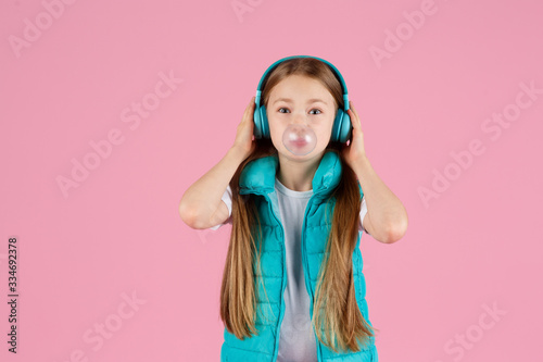 A little girl with headphones explodes pink chewing gum on a pink background