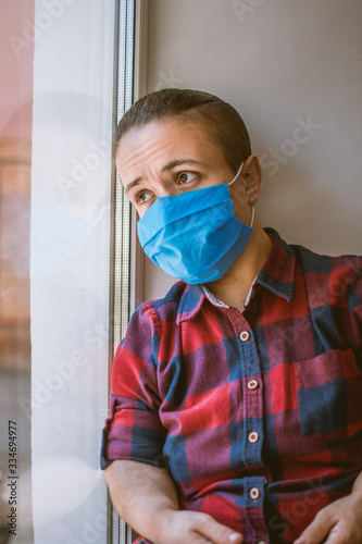 sad man in medical mask sits by the window of a house in isolation during a coronavirus pandemic