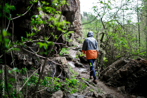 woman traveler in a gray-orange jacket with a hood and blue jeans walks along the rocks through the trees. outdoor activity concept