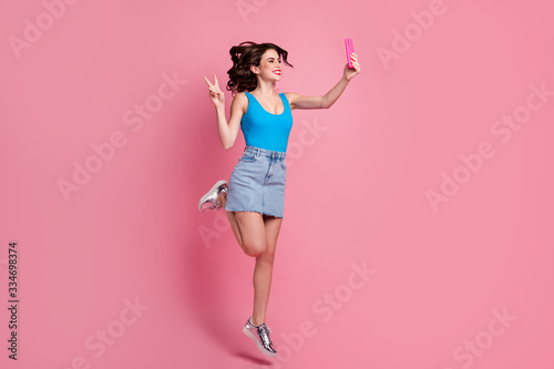 Full size profile photo of funny crazy lady jump high hold telephone making selfies show v-sign symbol wear blue tank-top denim short skirt shiny shoes isolated pink color background © deagreez