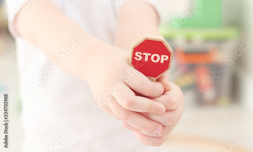 caucasian boy is clutching in palms and holding a close-up toy wooden road stop sign from the children's kit in a bright children's room. Teaching the road law, child safety concept, selective focus