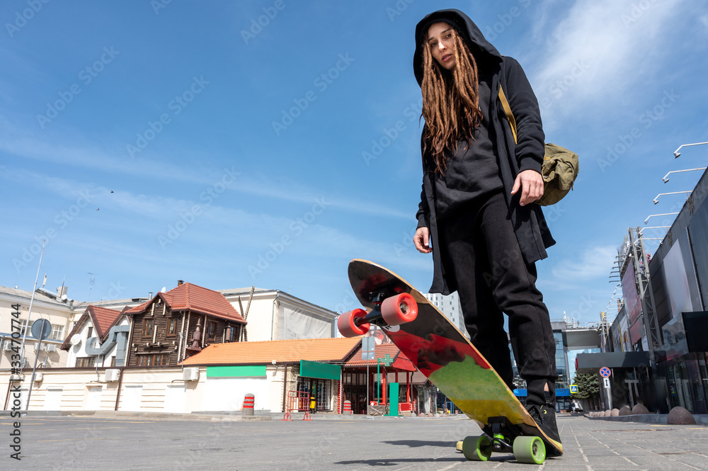 Naklejka premium girl with dreadlocks in a black hood and sneakers stands toe on a skateboard in the parking lot bottom view