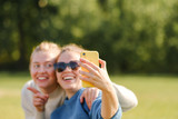 Young girls sit on the grass in the park and take a selfie on a smartphone. Girls enjoy the weekend outdoors. 