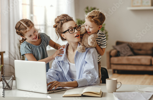 Little children distracting dedicated young woman working on laptop at home.