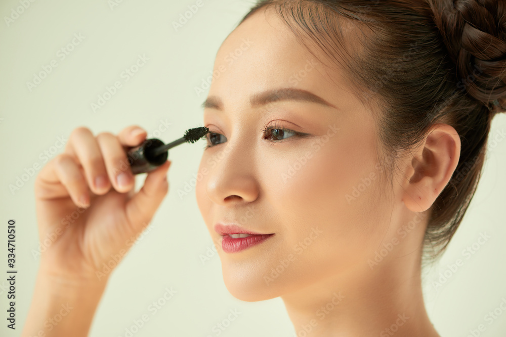 Close up portrait of young Asian woman applying mascara against light background. Close up portrait of young Asian woman applying mascara against light background.