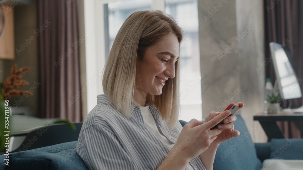 Caucasian woman sitting on couch in modern home using phone and smiling texting message watching contents or bloggs scrolling tapping apartment technology apps shopping sunlight sunny day