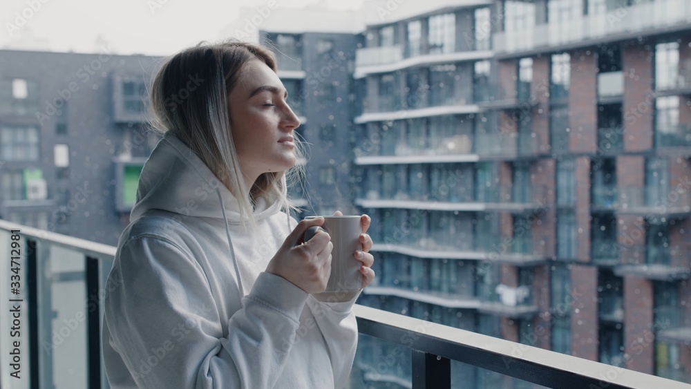 Attractive young woman in white sweater stays on balcony holds cup of hot coffee or tea watching a beautiful urban view and snow then close eyes and enjoy relax breathing fresh freezing air Attractive young woman in white sweater stays on balcony holds cup of hot coffee or tea watching a beautiful urban view and snow then close eyes and enjoy relax breathing fresh freezing air