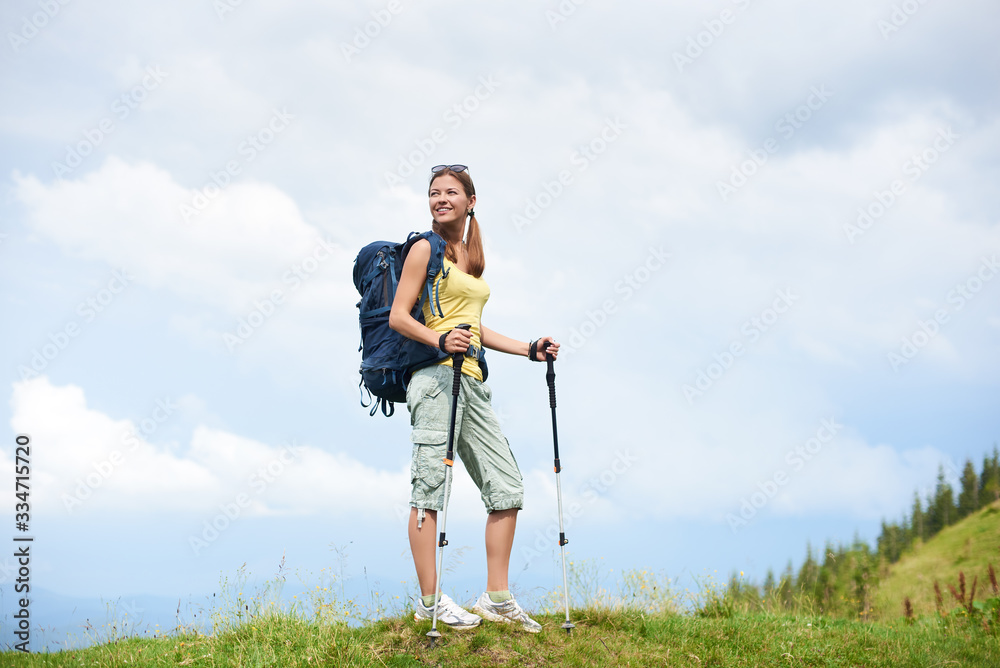 Beautiful happy woman hiker hiking mountain trail, walking on grassy hill, wearing backpack, using trekking sticks, enjoying summer cloudy day. Outdoor activity, tourism concept Beautiful happy woman hiker hiking mountain trail, walking on grassy hill, wearing backpack, using trekking sticks, enjoying summer cloudy day. Outdoor activity, tourism concept
