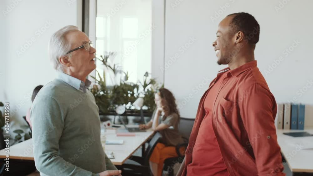 Cheerful senior caucasian man smiling, greeting young African American coworker with fist bumps and chatting with him while meeting together in office in the beginning of workday Cheerful senior caucasian man smiling, greeting young African American coworker with fist bumps and chatting with him while meeting together in office in the beginning of workday