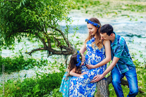 Pregnant woman mother to be in wreath and blue dress sitting with her daughter and husband near scenic river in sunset time in summer. Pregnancy and healthy organic close to nature lifestyle.