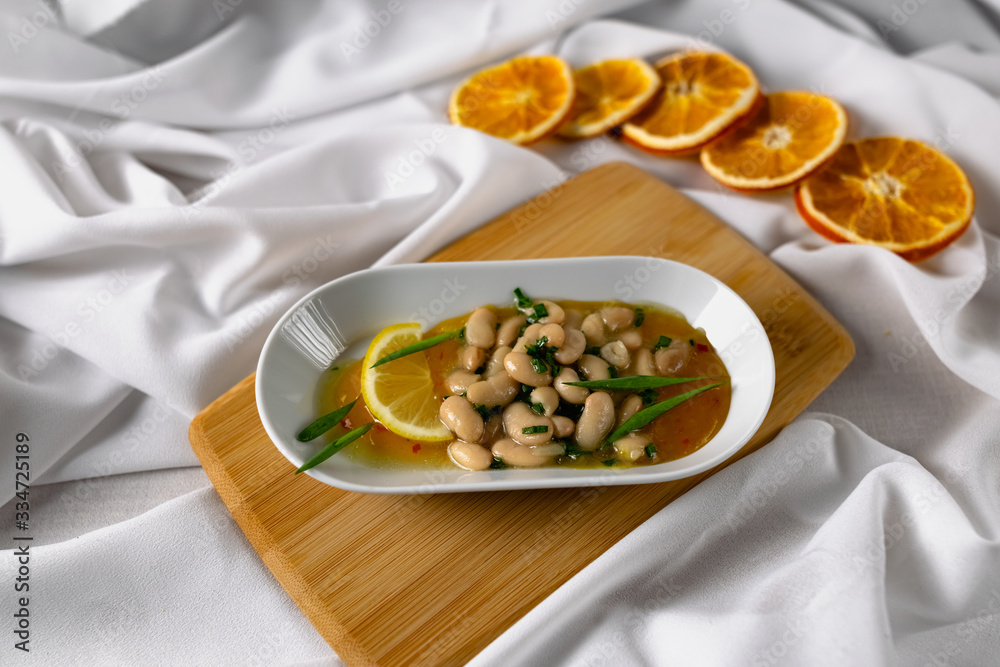 Marinated white beans in their own juice on an oval plate with Thai pineapple sauce and green onions. Decorated with a slice of lemon and dried oranges on a wooden Board with a snow white fabric