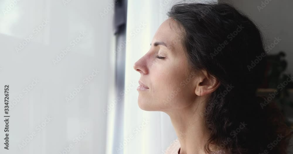 Calm serene young lady taking deep breath of fresh air standing at home window. Mindful healthy adult woman meditating with eyes closed at home feeling freedom. No stress breathing exercise concept