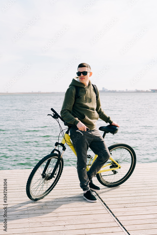 Fototapeta premium a young man in khaki sportswear and glasses stands on a pontoon near the lake and holds a yellow Bicycle near him on a Sunny spring day
