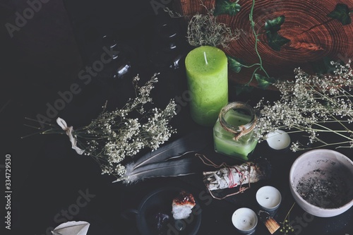 Various enchanted spell work ingredients on wiccan witch altar. Messy clutter on black table, with dried baby's breath flowers, green and white candles, dark grey feathers, crystals, sage sticks on it