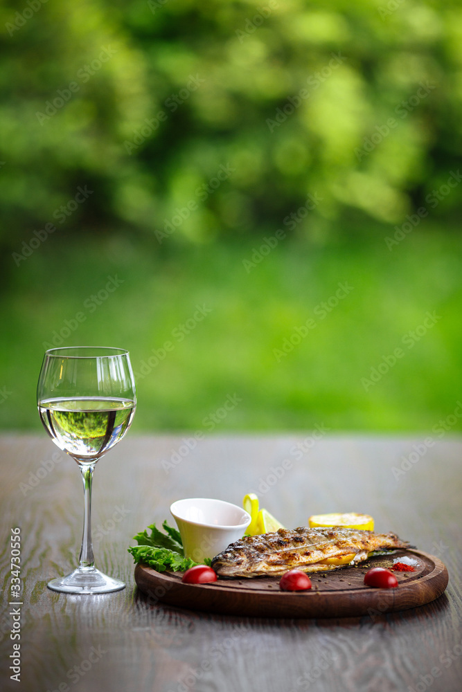 Roasted fish with vegetables on the wooden board with a glass of white wine on the wooden table, green background, vertical, side view Roasted fish with vegetables on the wooden board with a glass of white wine on the wooden table, green background, vertical, side view
