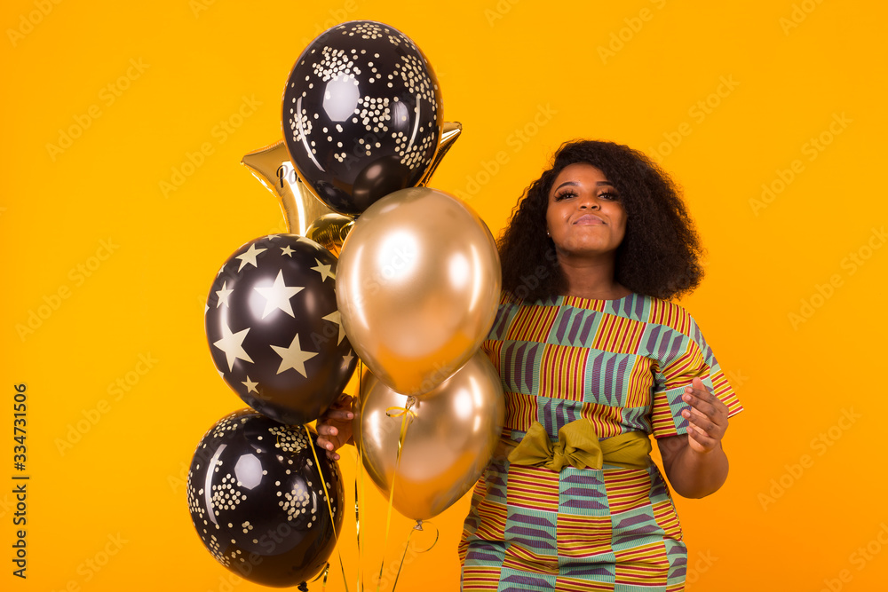 Naklejka premium Holidays, birthday party and fun concept - Portrait of smiling young African-American young woman looking sweet on yellow background holding balloons.