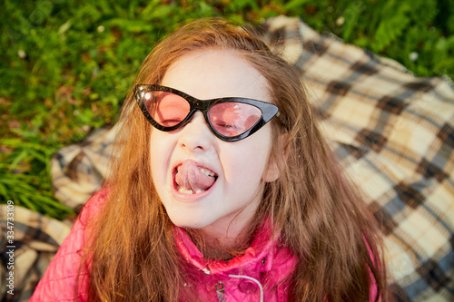 Playful funny little girl with long hair and in pink glasses having rest and fun on green grass in a summer park © keleny